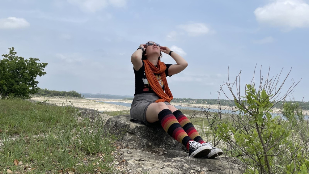 A person sits on a rock in the middle of open country looking up at the partial eclipse, holding eclipse glasses over their eyes with their hands