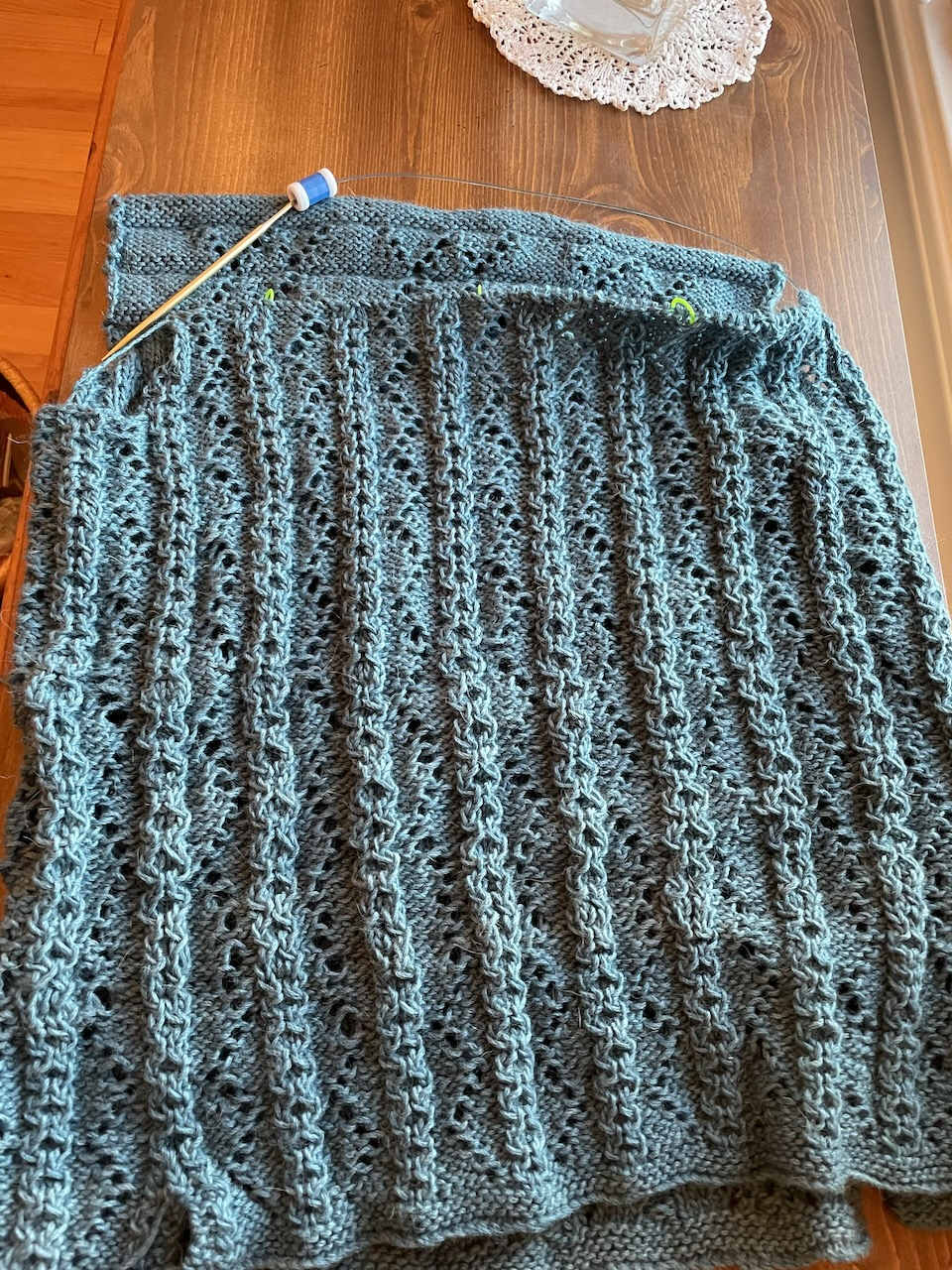 A blue sweater in progress with a complex lace and cable pattern. In the background, the edge of a knit doily.