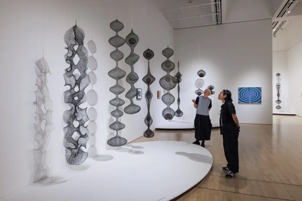 Two women observ hanging wire basket sculptures by Ruth Asawa in a gallery at the SFMOMA