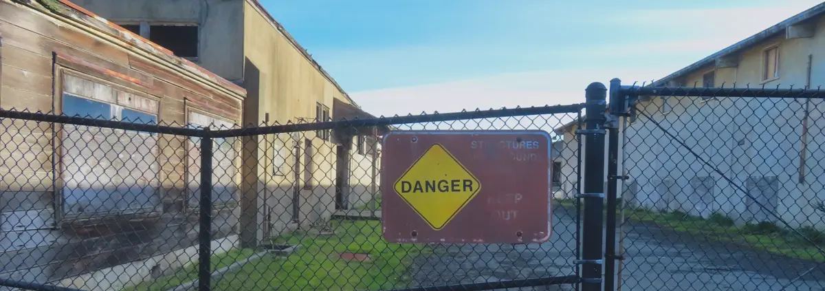 A photo of of a chain-link fence with a partly faded sign that says Danger and all the text about the nature of the danger is illegible. Behind the chain-link fence, decaying buildings and the sky.