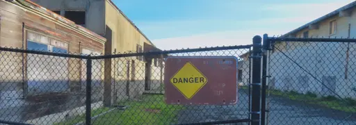 A photo of of a chain-link fence with a partly faded sign that says Danger and all the text about the nature of the danger is illegible. Behind the chain-link fence, decaying buildings and the sky.