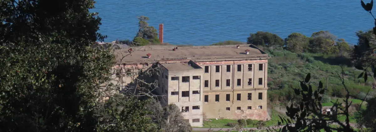 A photo of an abandoned building with empty windows seen from above among trees and coastal chaparral with blue ocean behind it.
