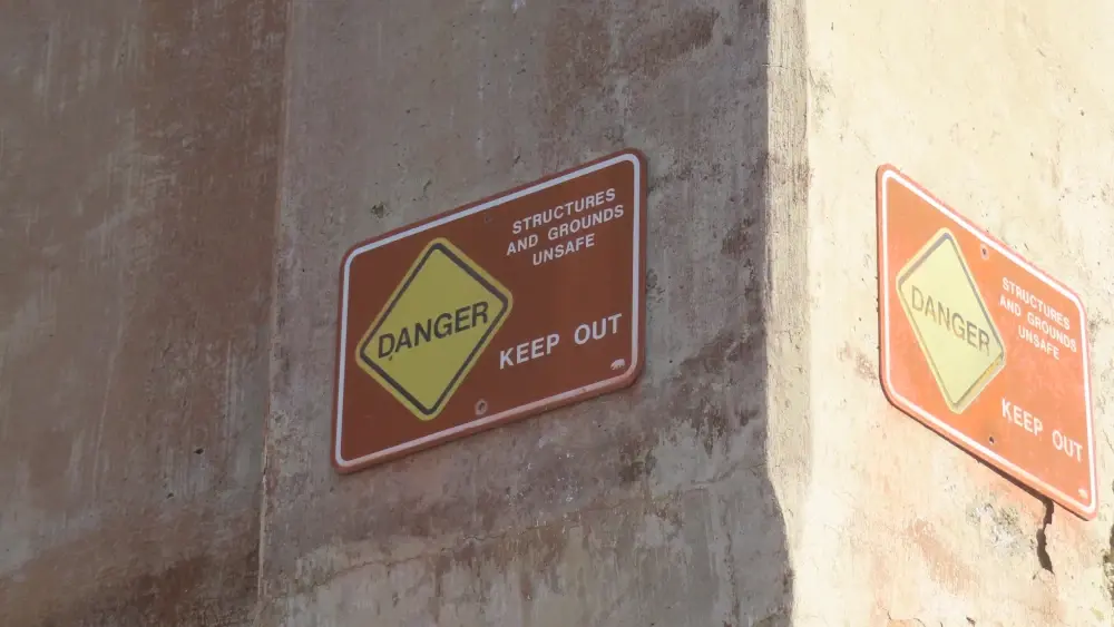 A warning sign on a the corner of a building that says DANGER STRUCTURES AND GROUNDS UNSAFE KEEP OUT
