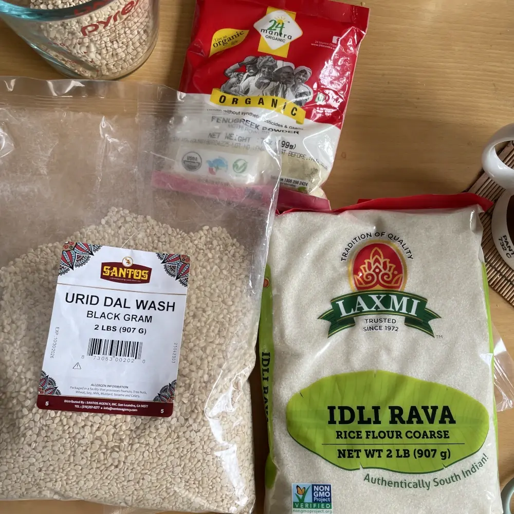 Three bags of dry food lie flat on a table: urad dal, idli rava, and fenugreek. Also a glass measuring cup with some of the dal.