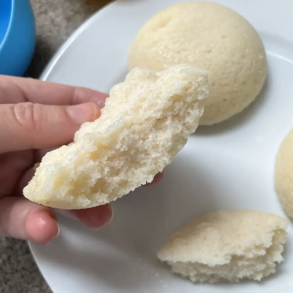 An idli broken in half, showing the inside crumb which is fluffy. However the top of the idli is not very risen at all.
