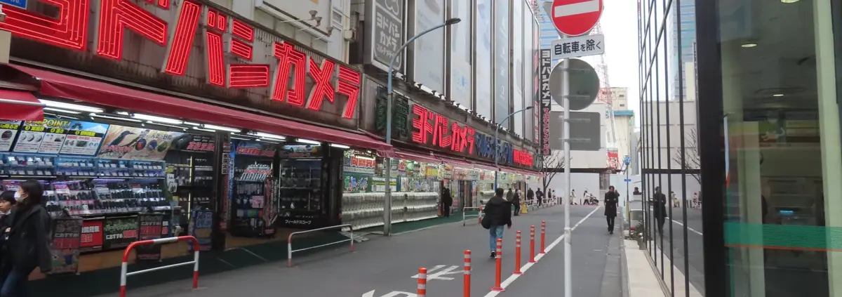 View into a side-street in Tokyo. A red neon sign in characters stretches length-wise over multiple storefronts displaying electronics and then rows and rows of gacha machines. On the right, the bottom of a glass skyscraper is visible, and in the distance more city to infinity.