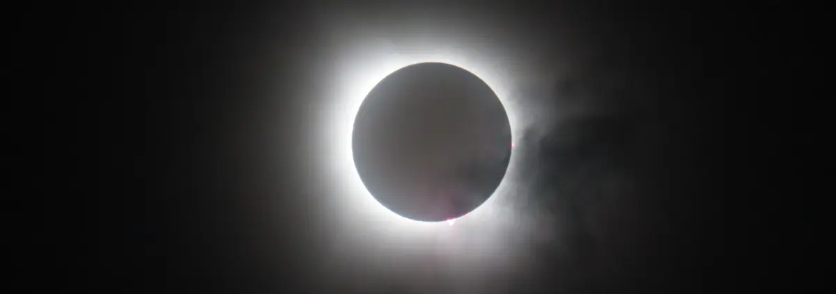 A black hole in a black sky surrounded by a white corona and spots of red around the disk of the black sun.