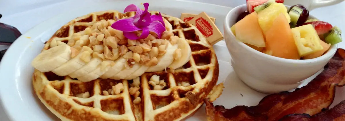 A photo of a waffle topped with a sliced banana and nuts and decorated with a purple orchid flower rests on a plate. To the side stands a small bowl of fruit salad with of a slice of fried American bacon next to it.