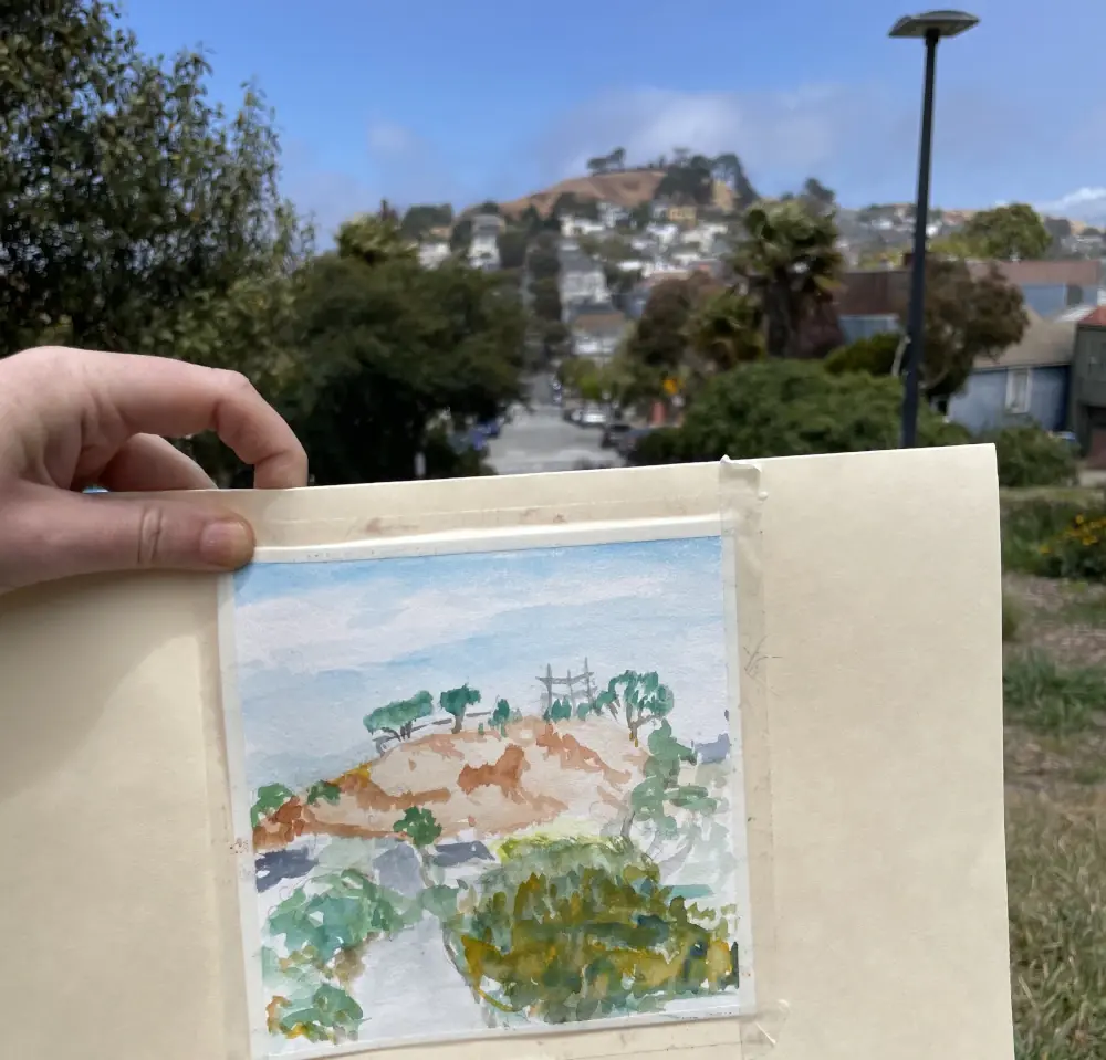 A watercolor sketch of Bernal Hill held up next to the view of the same hill