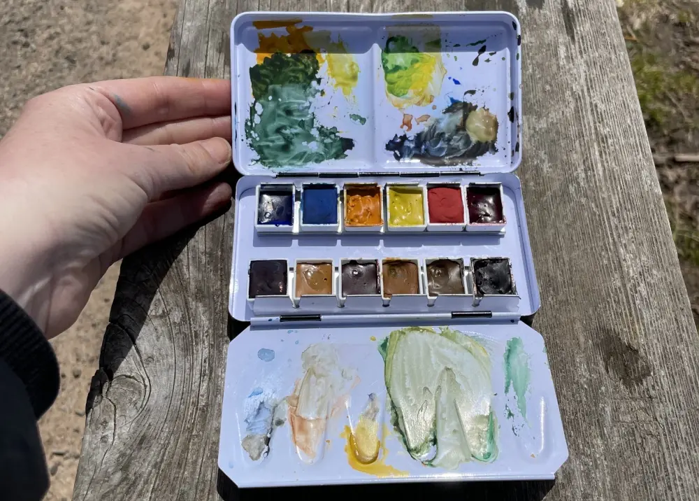A messy watercolor palette with 12 half pans of paint in the middle and splotches of mixed paint on the foldout palette above and below