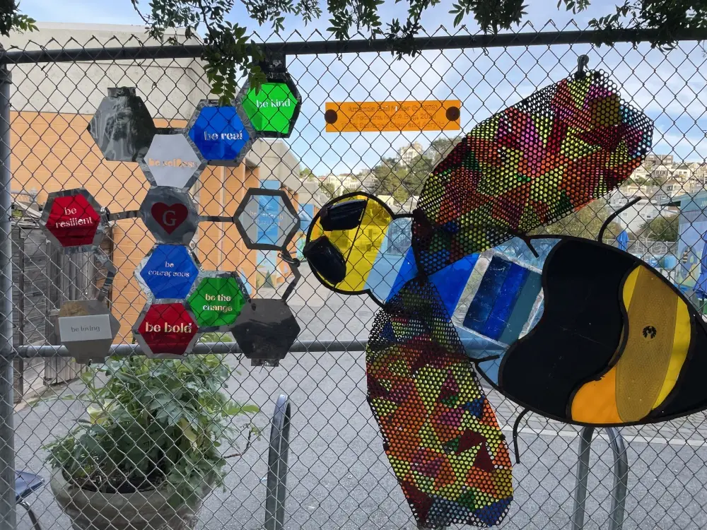 A flat plastic bee attached to a chain link fence faces a series of colorful hexagons with inspirational messages including
