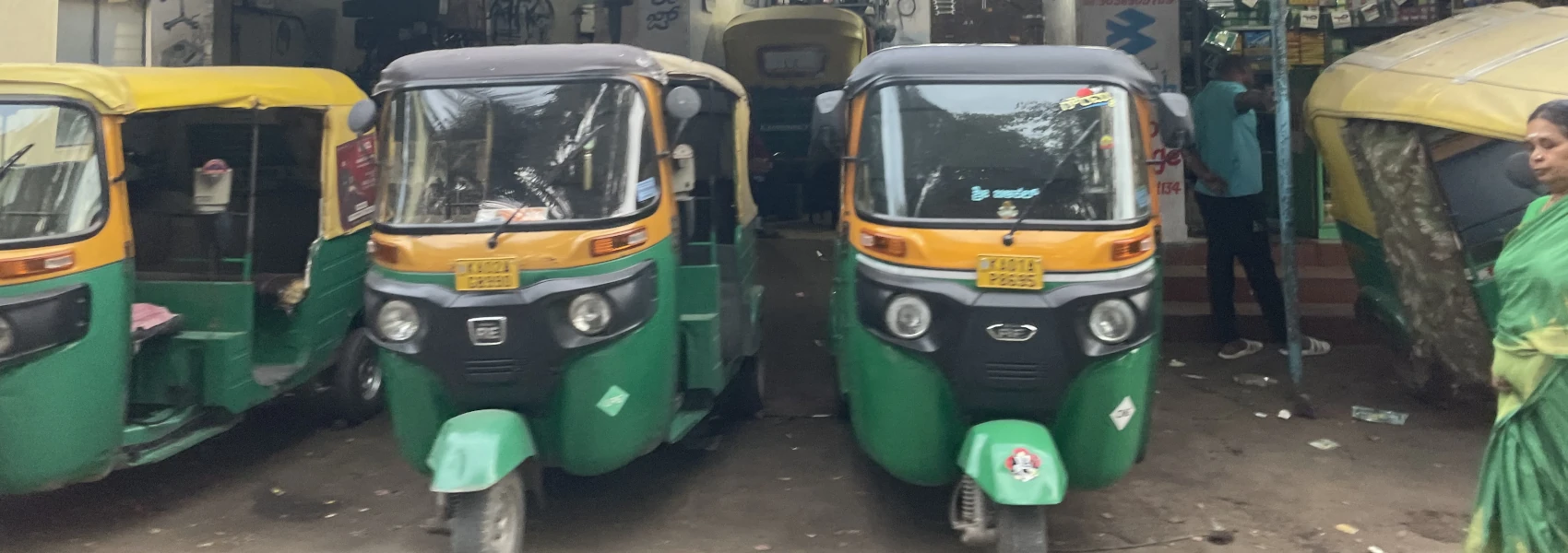 Three green and yellow autos, also known as tuk-tuks, parked in front of a shop that services them. Another auto can be seen inside. A lady wearing a sari in the same shade of green and yellow is just entering the frame on the right.