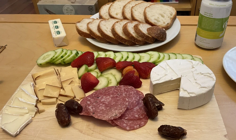 A charcuterie board with cucumber slices, strawberries, smoked gouda, brie, and dates. Sliced bread in the background.