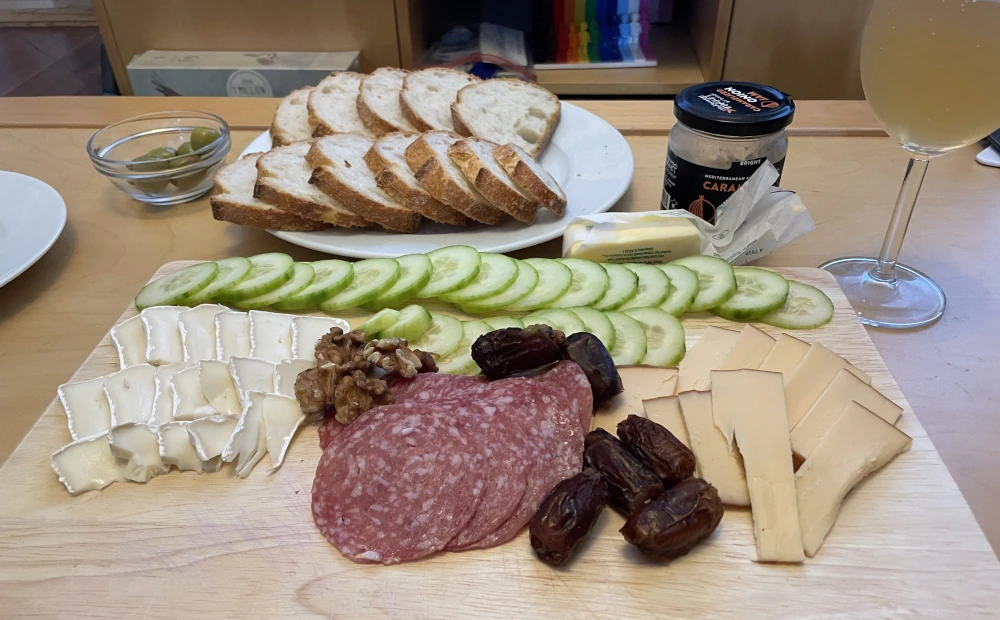 A charcuterie board with cucumber slices, brie, walnuts, salami, dates, and smoked gouda. A glass of amber liquid stands to one side, a little bowl of olives, and a plate of bread behind.