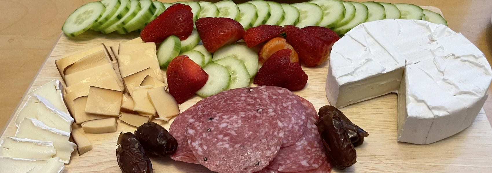 Charcuterie board with cut cucumber, halved strawberries, a wheel of brie, salami slices, dried dates