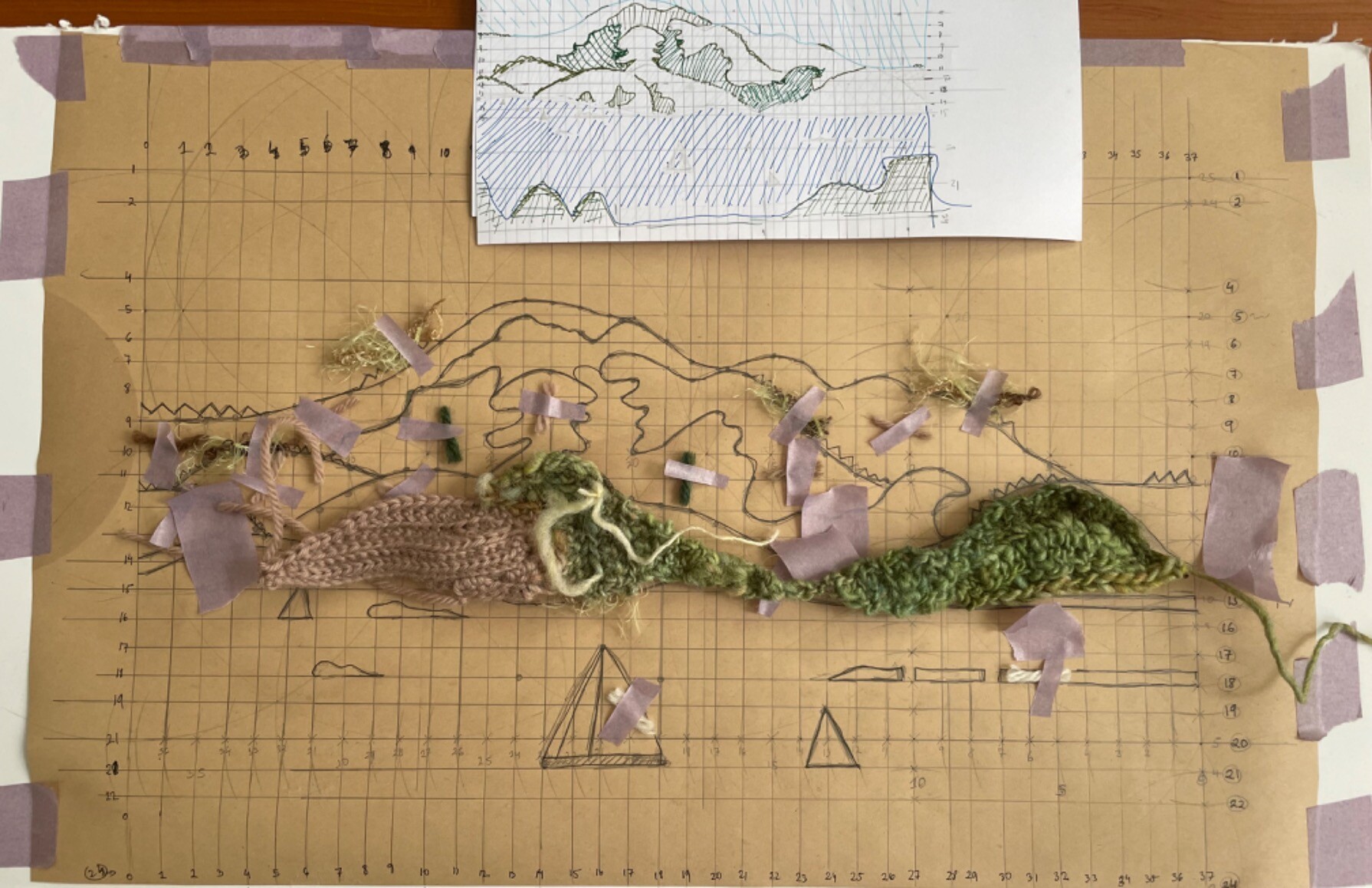 Butcher’s paper with a grid drawn on it, and a diagram of an island. Bits of yarn are taped to portions. A little bit of the bottom part of the island is filled in with messy crochet.