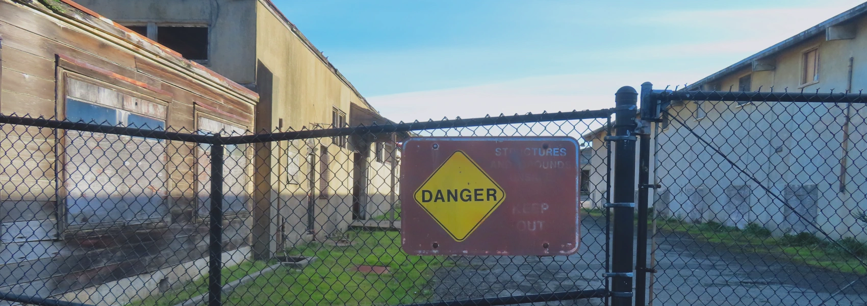 A photo of of a chain-link fence with a partly faded sign that says Danger and all the text about the nature of the danger is illegible. Behind the chain-link fence, decaying buildings and the sky.