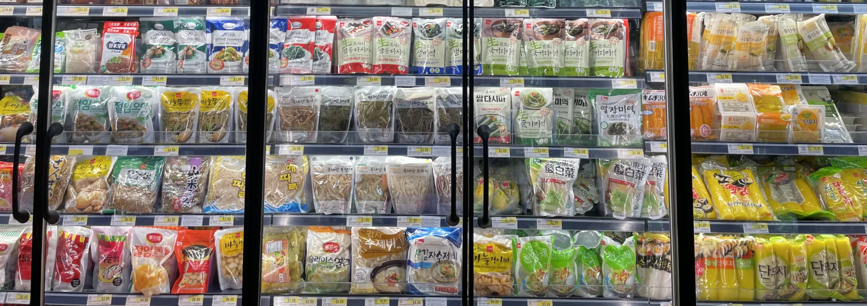 A cropped view of a glass-fronted fridge cabinet displaying a variety of packaged pickled vegetables, including seaweed and radish.