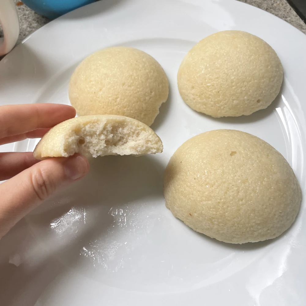 Three idli on a plate. The fourth is held up in a hand and broken in half showing a dense crumb inside