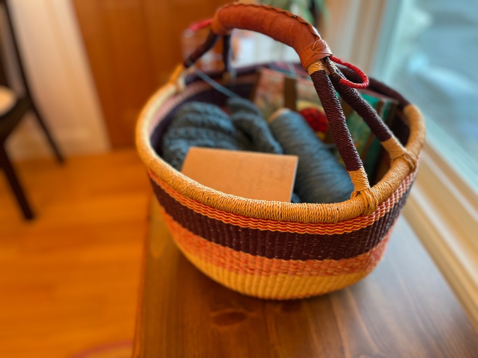 A round basket with a handle. Inside is a spool of blue yarn, some bit of knitted fabric, and a small notebook.