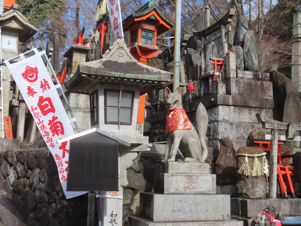 A stone fox seems to grin at the viewer amids a multitude of shrines and and lanterns and rice straw chanins