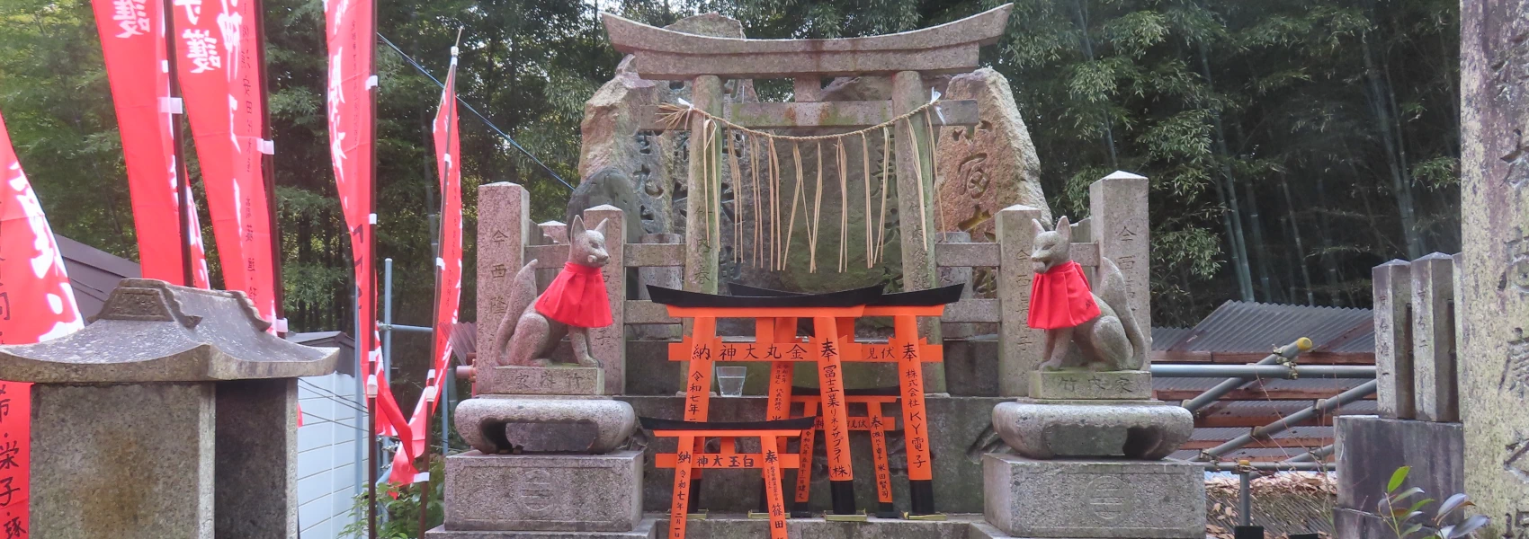 A stone alar framed by two stone foxes wearing bibs. A straw chain decorates the stone torii behind the foxes.