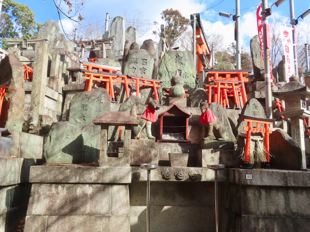 Small and medium stone shrines with Japanese writing on them, small red torii leaning against them, and a pair of stone foxes wearing red bibs