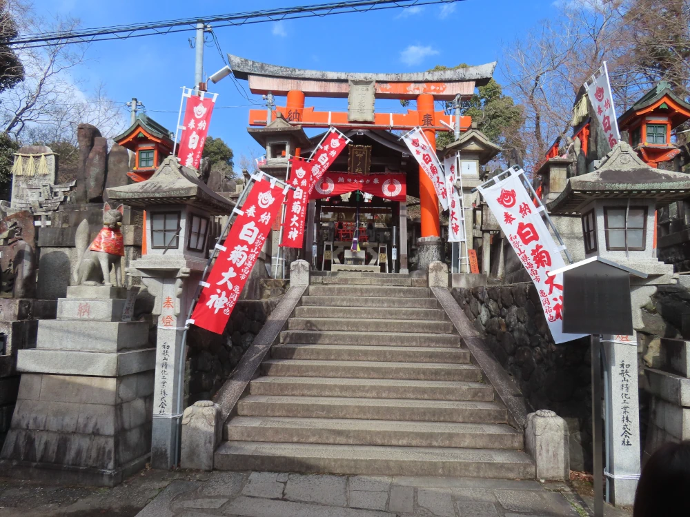 Stone stairs leading up to a red torii entrance to a more formal altar. It's framed by red and white flags and guarded by stone foxes