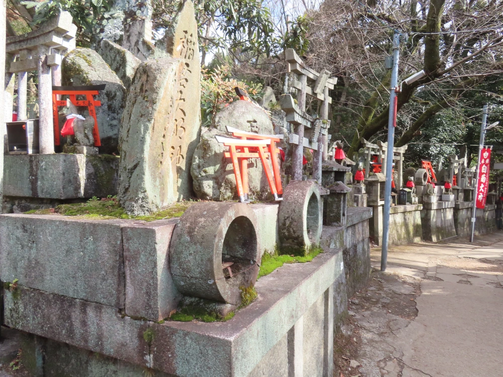 A side on view of a series of small stone altars with standing stones and stone torii and small red torii leaning against them