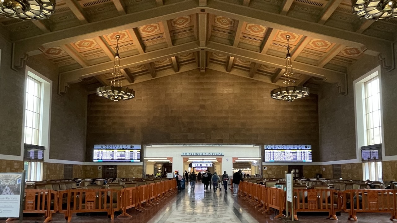 A highly decorative waiting room in Union Station with a decorative floor, wooden beam ceilings with elaborate chandeliers, but the seats are blocked off by garish orange barriers to prevent anyone from sitting there who doesn't have a ticket.