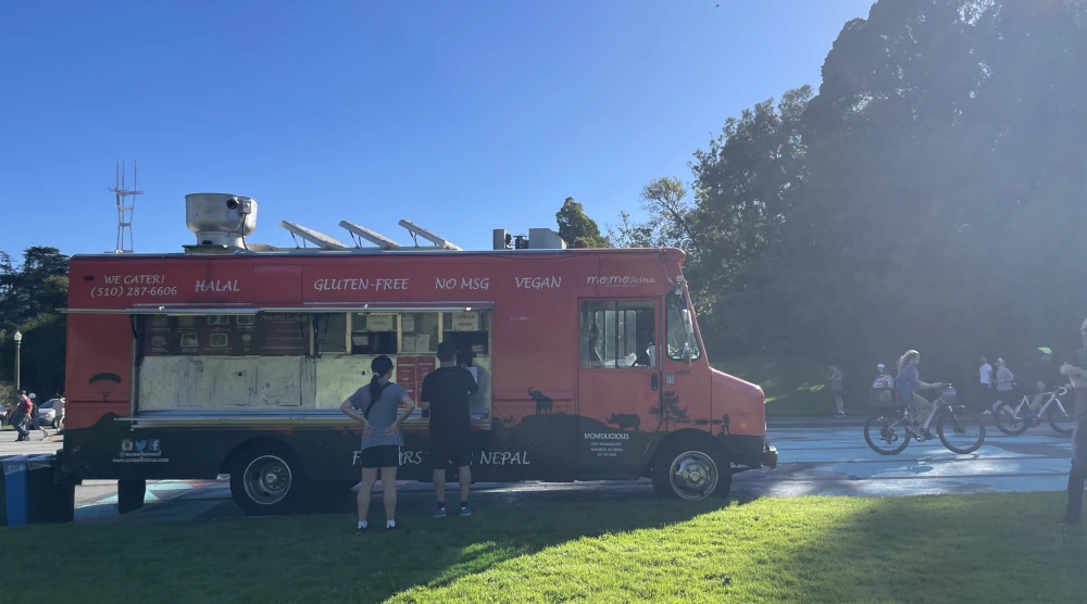 A red food truck selling momos, signs on it advertise that they're halal, vegan and guletn free. Two people stand in front of it ordering. In the background, bikers zoom by on the road.