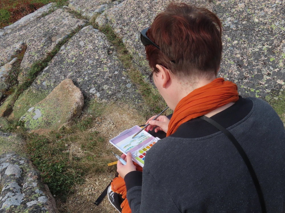 A person bends over a small watercolor palette while sitting on some rocks