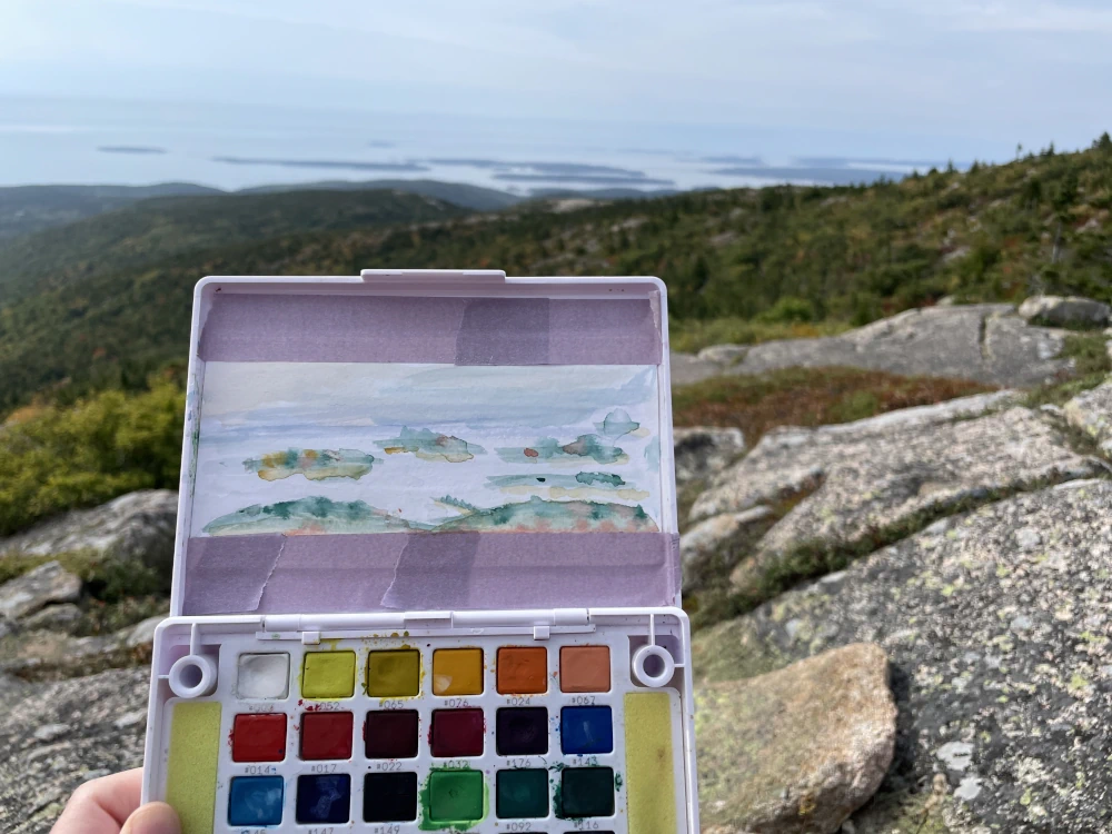 A photo of a portable watercolor palette with a painting of a scene of coastal islands it depicts