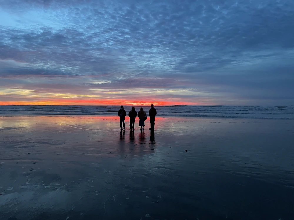 Four human figures silhouetted before a red streak of sunset sky, a line of ocean waves, and the dappled cloudy sky and its reflection in the wet beach sand. Layers of color with horizontal symmetry.