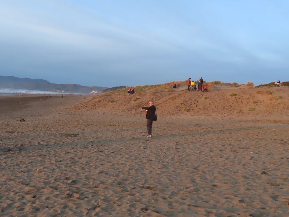 A man in black standing against golden hour lit dunes holds up his phone to take a photo.