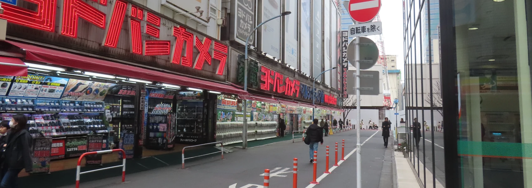 View into a side-street in Tokyo. A red neon sign in characters stretches length-wise over multiple storefronts displaying electronics and then rows and rows of gacha machines. On the right, the bottom of a glass skyscraper is visible, and in the distance more city to infinity.