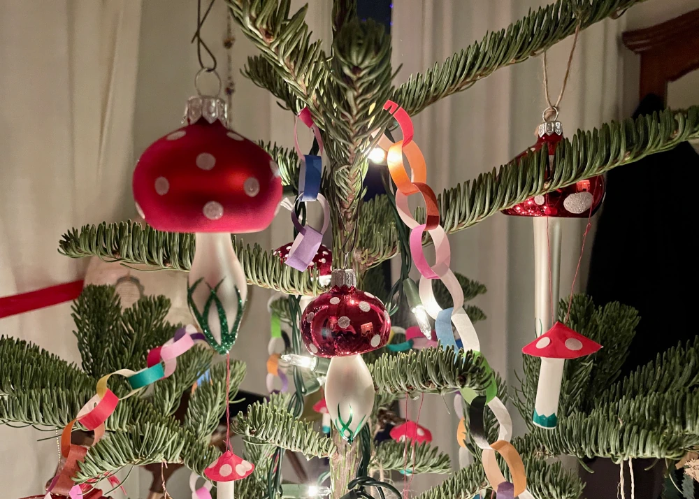 A glass mushroom bauble is in the foreground. It has red cap and wite spots, and the stem is white with sparky green lines at the base representing grass. There are several more mushroom baubles in the background hanging on a Christmas tree.