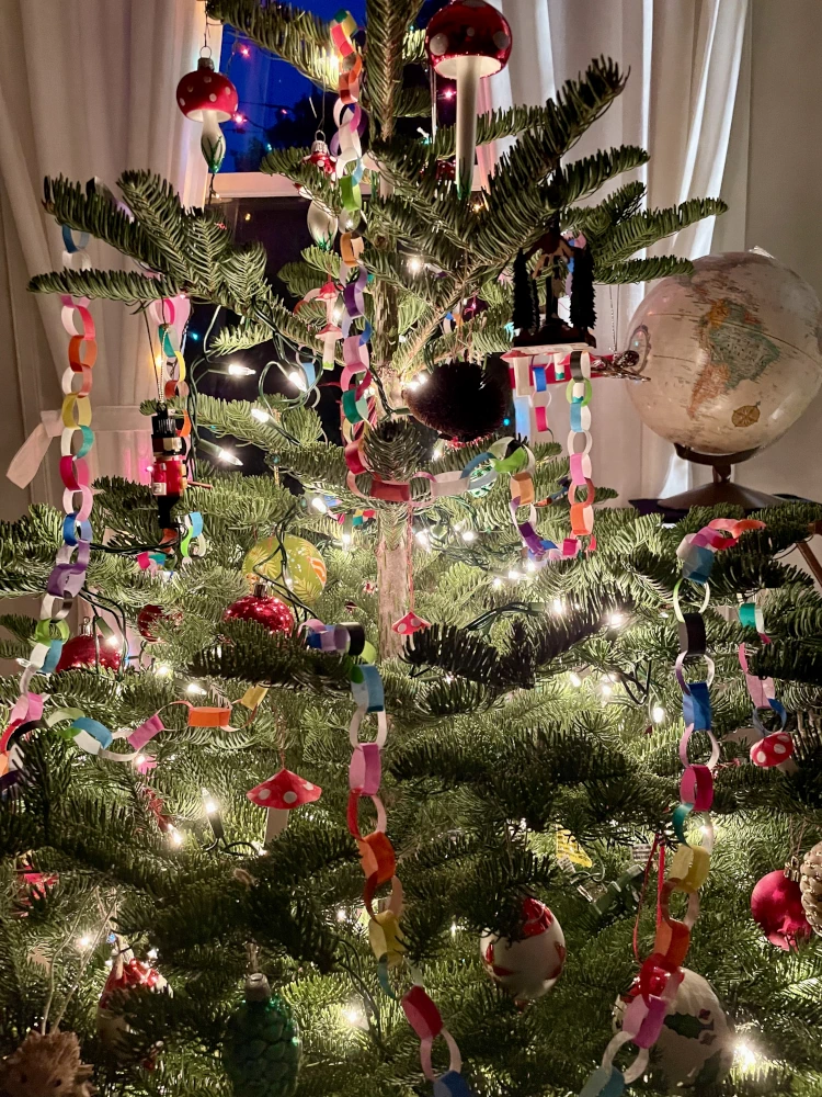 A Christmas tree decorated with colorful paper chains, white lights, red baubles, paper mushrooms, and glass mushrooms.